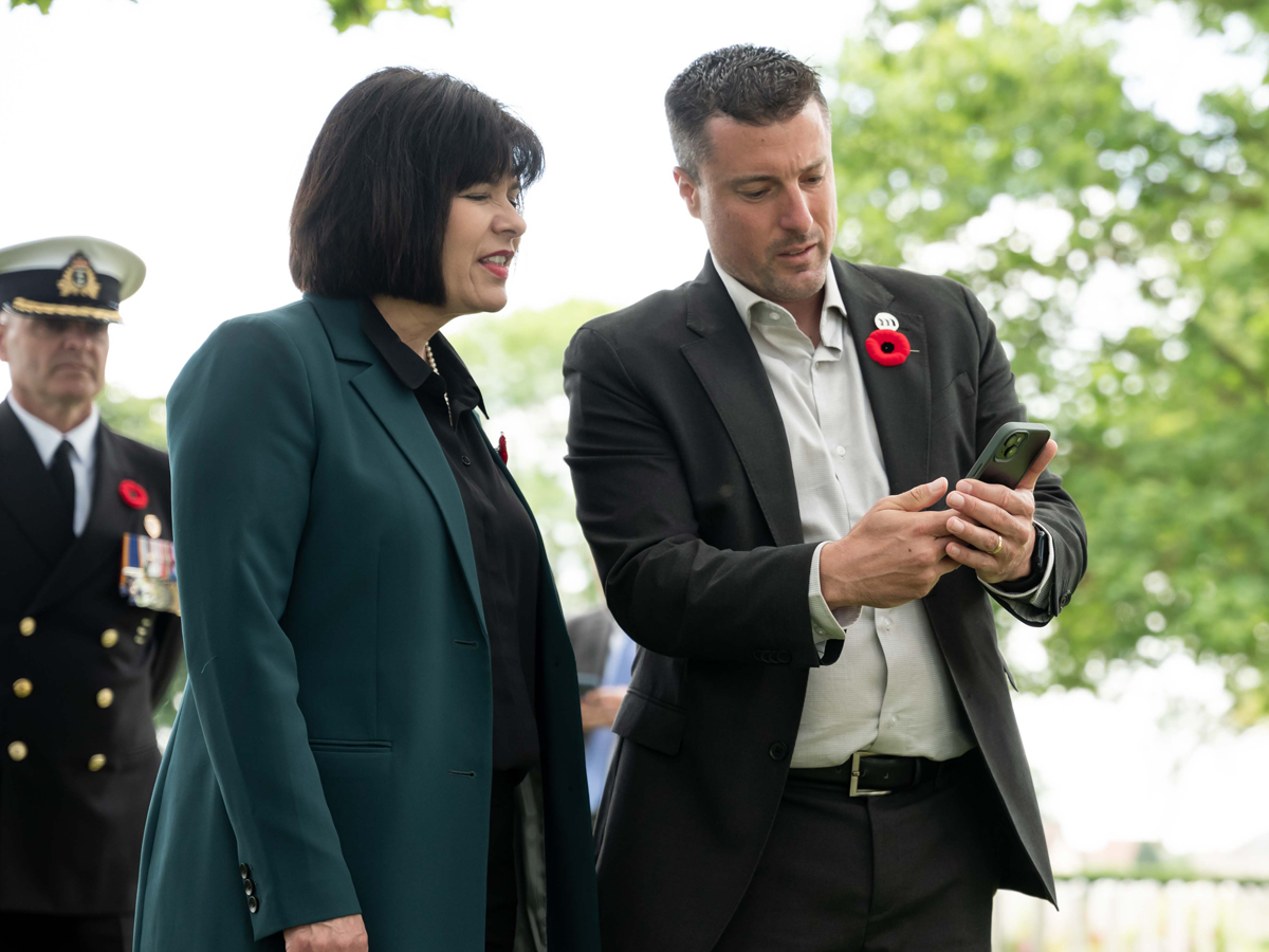 La ministre des Anciens Combattants et ministre associée de la Défense nationale, Ginette Petitpas Taylor, à gauche, et le cofondateur de l'Ancre de la mémoire, Ryan Mullens, utilisent l'application interactive de l'entreprise pendant les services du jour J 80 au cimetière de guerre canadien de Bretteville-sur-Laize, en Normandie, en France. Photo : Anciens Combattants Canada : Anciens Combattants Canada